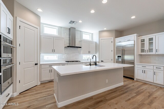 kitchen with appliances with stainless steel finishes, light hardwood / wood-style flooring, a center island with sink, sink, and wall chimney exhaust hood