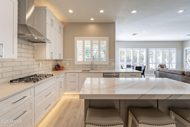 kitchen featuring wall chimney range hood, light stone countertops, stainless steel appliances, a sink, and a kitchen breakfast bar