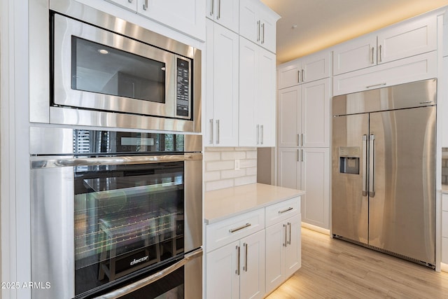 kitchen featuring light wood finished floors, backsplash, built in appliances, light countertops, and white cabinetry