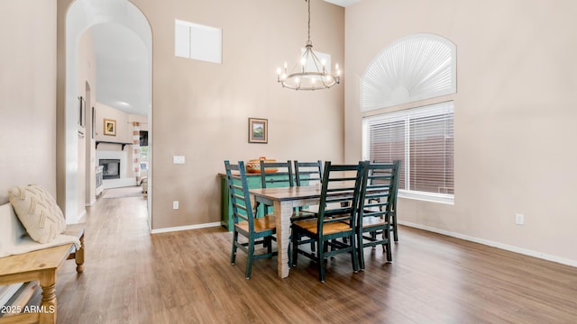 dining space with a towering ceiling, wood finished floors, a chandelier, baseboards, and a glass covered fireplace