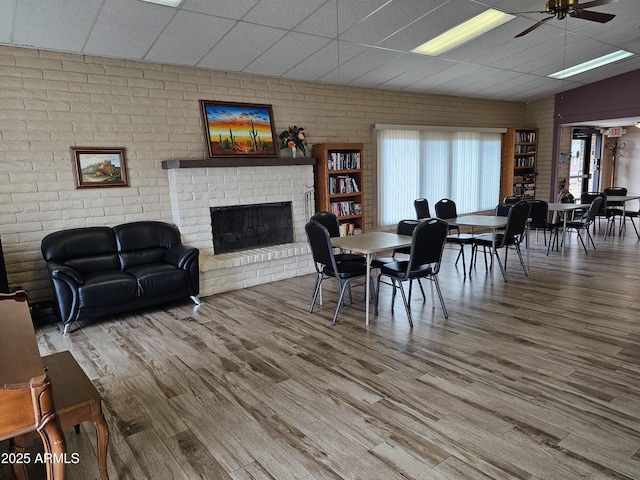 dining space featuring a brick fireplace, brick wall, a drop ceiling, wood finished floors, and a ceiling fan