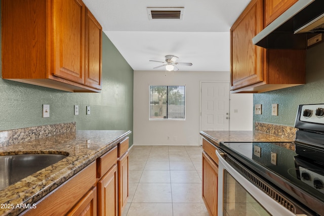 kitchen featuring stainless steel range with electric stovetop, under cabinet range hood, dark stone counters, ceiling fan, and brown cabinetry