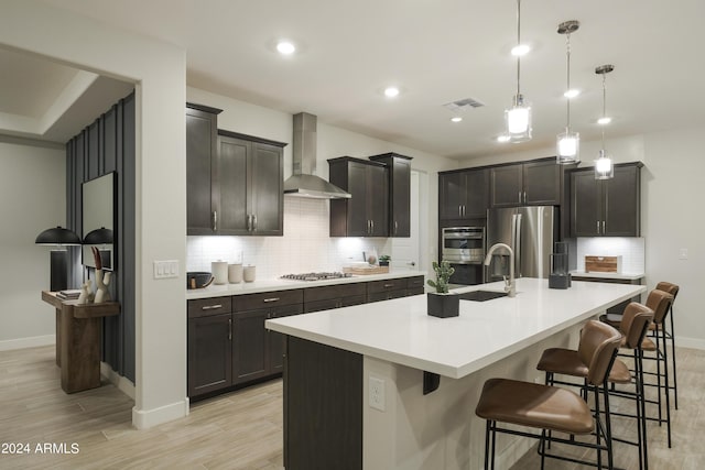 kitchen featuring a breakfast bar, hanging light fixtures, wall chimney exhaust hood, an island with sink, and appliances with stainless steel finishes