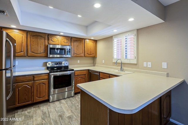 kitchen featuring a raised ceiling, light wood-style flooring, appliances with stainless steel finishes, a peninsula, and a sink