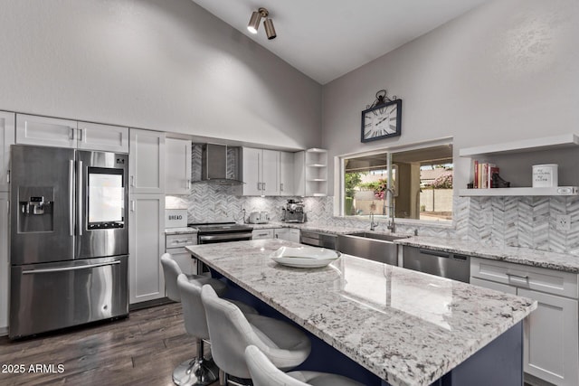 kitchen with high vaulted ceiling, open shelves, wall chimney exhaust hood, appliances with stainless steel finishes, and a kitchen island