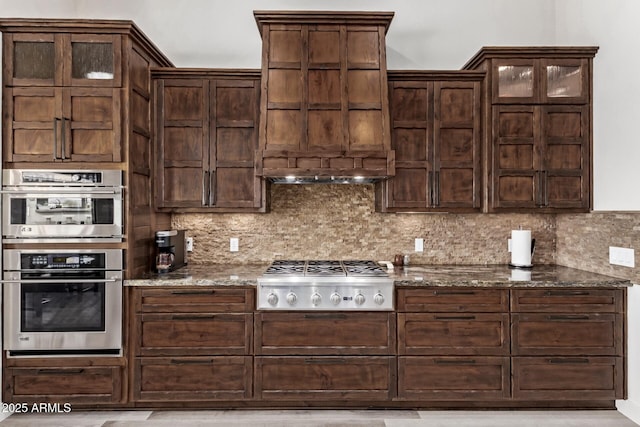 kitchen with backsplash, premium range hood, dark stone countertops, dark brown cabinetry, and stainless steel appliances