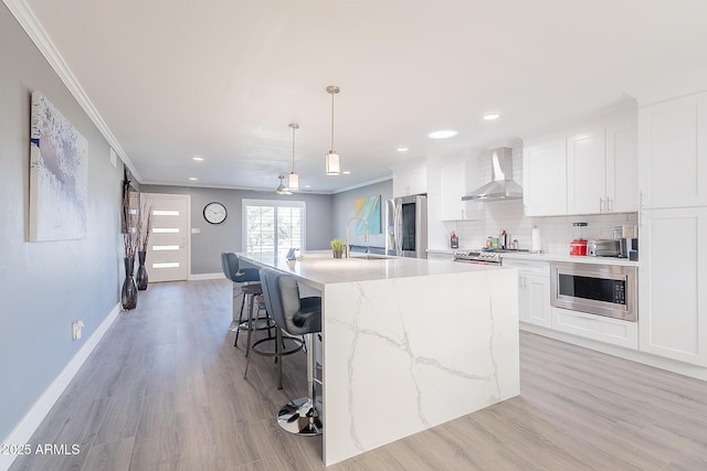 kitchen with white cabinets, hanging light fixtures, stainless steel appliances, a center island with sink, and wall chimney exhaust hood