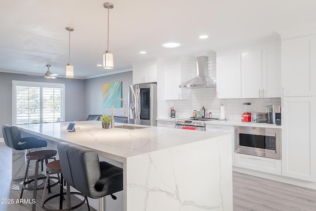 kitchen with wall chimney range hood, sink, a kitchen island with sink, white cabinetry, and stainless steel appliances