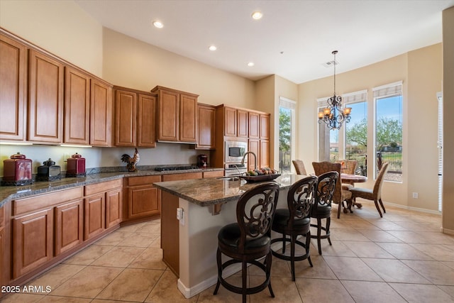 kitchen featuring light tile patterned flooring, a kitchen breakfast bar, stainless steel microwave, an inviting chandelier, and brown cabinetry