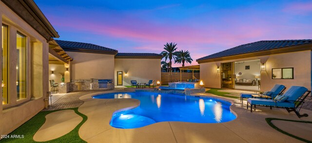 pool at dusk featuring a pool with connected hot tub, fence, exterior kitchen, and a patio