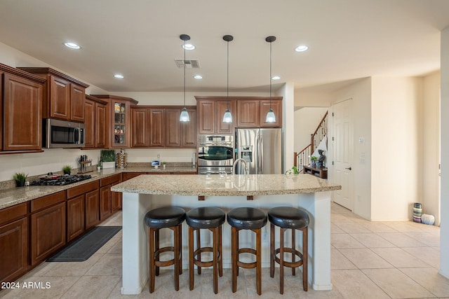 kitchen featuring appliances with stainless steel finishes, glass insert cabinets, hanging light fixtures, light stone countertops, and a kitchen island with sink