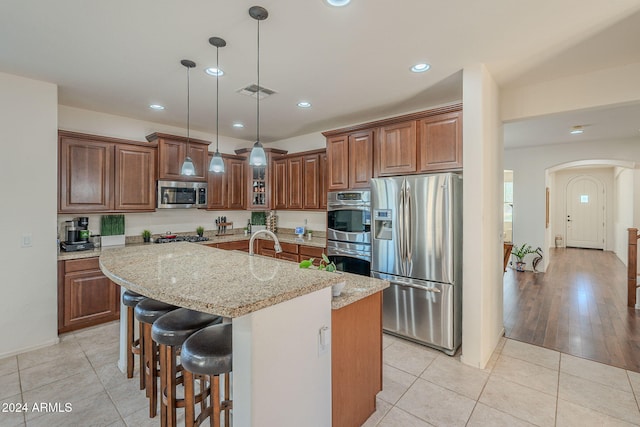 kitchen with light stone counters, stainless steel appliances, a kitchen island with sink, decorative light fixtures, and light tile patterned flooring