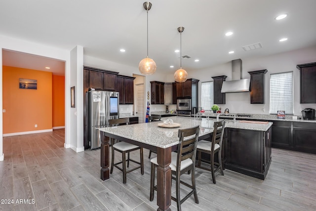 kitchen featuring wall chimney range hood, light hardwood / wood-style floors, decorative light fixtures, a kitchen island, and appliances with stainless steel finishes