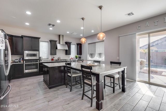 kitchen with a center island, wall chimney exhaust hood, light hardwood / wood-style flooring, pendant lighting, and appliances with stainless steel finishes