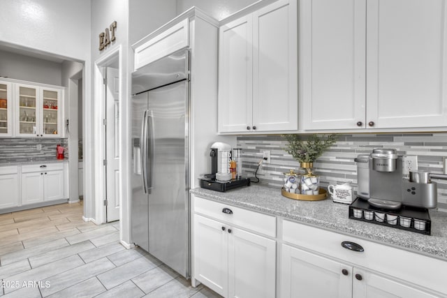 kitchen featuring light stone counters, white cabinets, glass insert cabinets, stainless steel built in fridge, and backsplash
