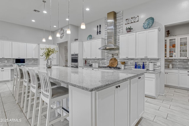 kitchen with wall chimney exhaust hood, visible vents, backsplash, a large island with sink, and white cabinets