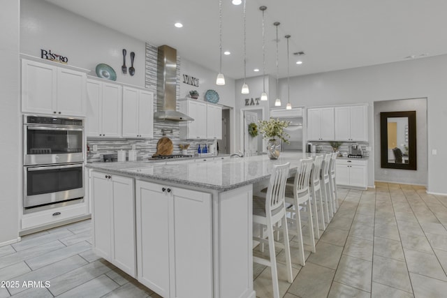 kitchen with wall chimney range hood, white cabinetry, decorative backsplash, appliances with stainless steel finishes, and a large island with sink