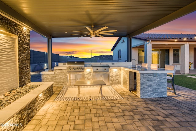 patio terrace at dusk with a sink, area for grilling, outdoor wet bar, a grill, and a ceiling fan