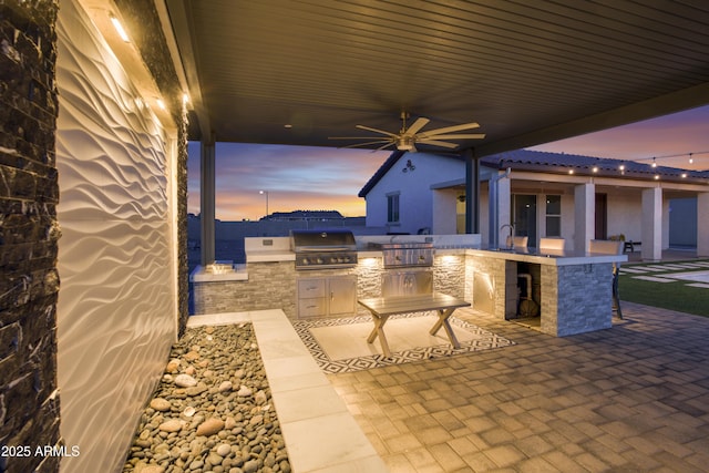 patio terrace at dusk featuring ceiling fan, a grill, and exterior kitchen