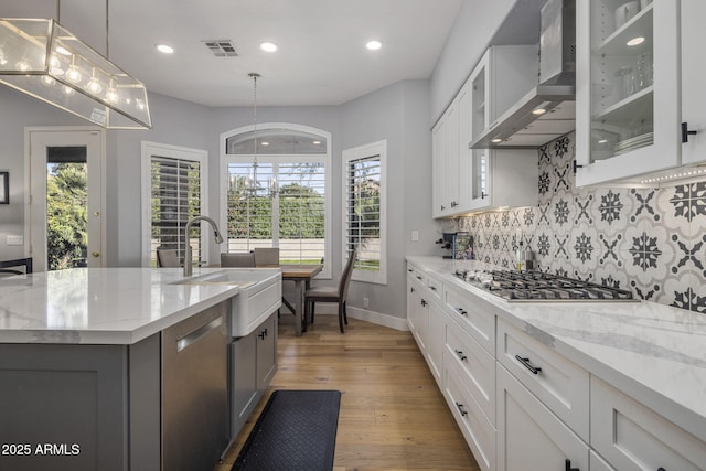 kitchen with wall chimney exhaust hood, white cabinetry, decorative light fixtures, stainless steel appliances, and a kitchen island with sink