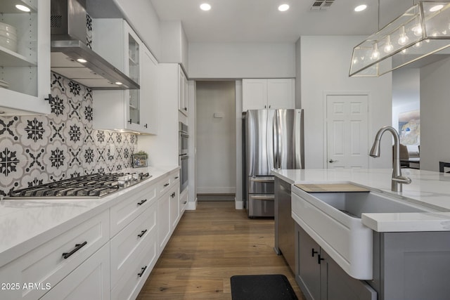 kitchen with sink, appliances with stainless steel finishes, white cabinetry, hanging light fixtures, and wall chimney exhaust hood