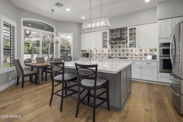 kitchen with pendant lighting, appliances with stainless steel finishes, white cabinetry, a kitchen island with sink, and wall chimney exhaust hood