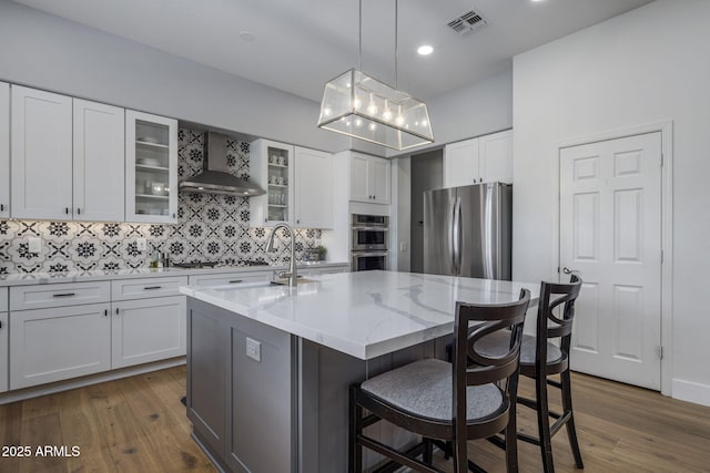 kitchen with appliances with stainless steel finishes, an island with sink, white cabinets, light stone countertops, and wall chimney range hood