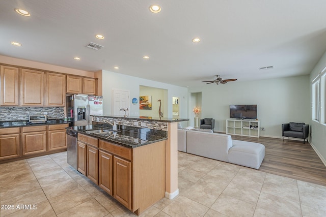kitchen featuring an island with sink, a ceiling fan, tasteful backsplash, stainless steel appliances, and dark stone counters