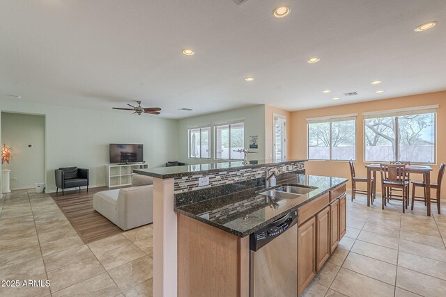 kitchen with light tile patterned floors, dark stone countertops, stainless steel dishwasher, a sink, and a kitchen island with sink