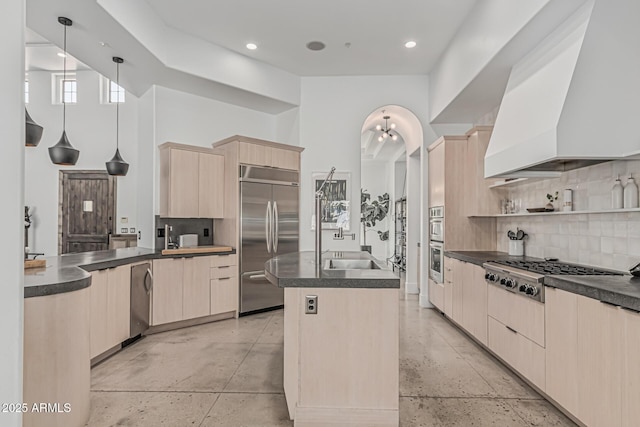 kitchen featuring light brown cabinetry, dark countertops, appliances with stainless steel finishes, a kitchen island with sink, and tasteful backsplash