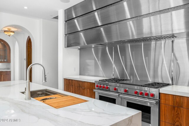 kitchen featuring wall chimney exhaust hood, light stone countertops, sink, and range with two ovens