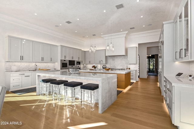 kitchen with a kitchen island with sink, white cabinets, decorative light fixtures, light wood-type flooring, and tasteful backsplash