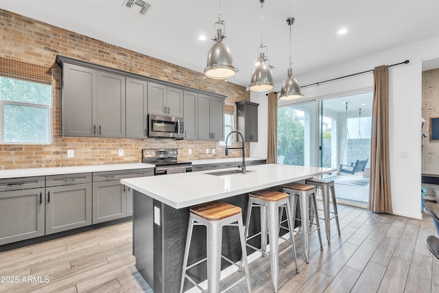 kitchen with a kitchen island with sink, hanging light fixtures, gray cabinetry, stainless steel appliances, and a kitchen breakfast bar