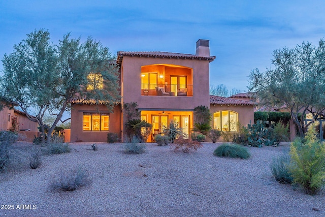 back of house at dusk with a chimney, a tile roof, a balcony, and stucco siding