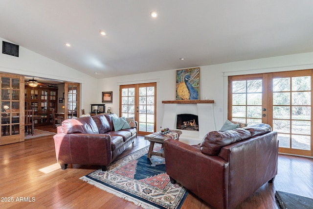 living area with french doors, visible vents, vaulted ceiling, wood finished floors, and a warm lit fireplace