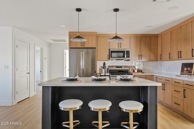 kitchen featuring stainless steel appliances, light hardwood / wood-style floors, light brown cabinetry, hanging light fixtures, and a center island with sink