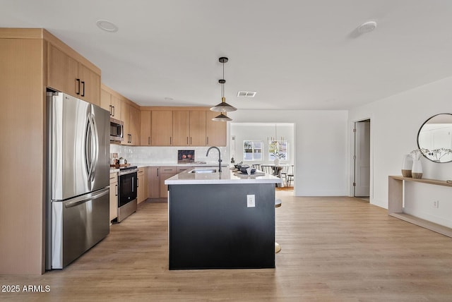 kitchen with stainless steel appliances, sink, light brown cabinets, an island with sink, and hanging light fixtures