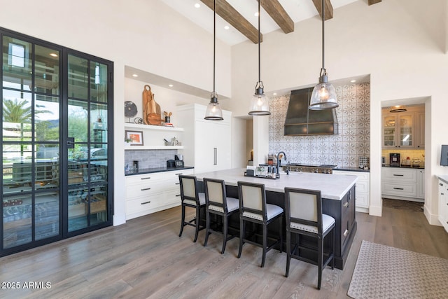 kitchen with pendant lighting, beamed ceiling, an island with sink, white cabinets, and custom exhaust hood