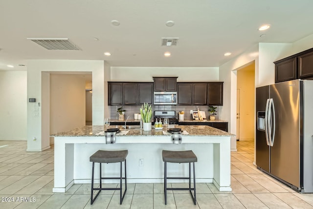 kitchen with dark brown cabinetry, tasteful backsplash, light stone counters, a kitchen island with sink, and appliances with stainless steel finishes