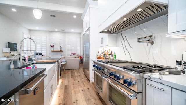 kitchen featuring decorative light fixtures, white cabinets, high end appliances, wall chimney range hood, and light wood-type flooring