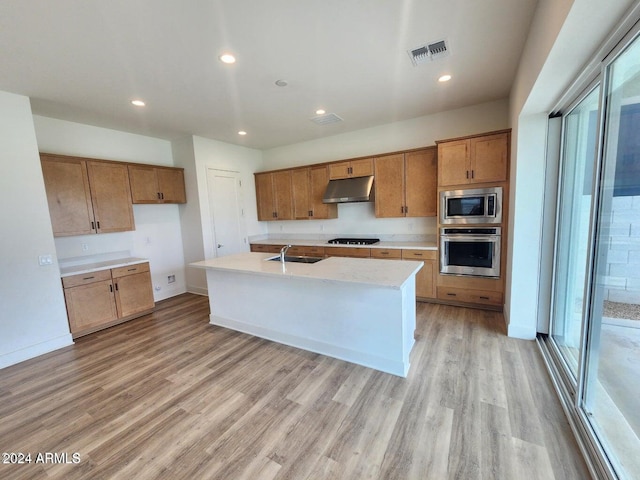 kitchen with sink, light hardwood / wood-style flooring, appliances with stainless steel finishes, a kitchen island with sink, and light stone counters