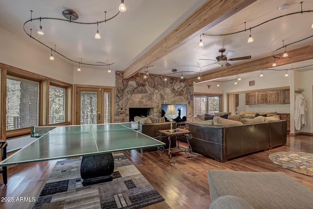 recreation room featuring rail lighting, a ceiling fan, a stone fireplace, beamed ceiling, and hardwood / wood-style flooring