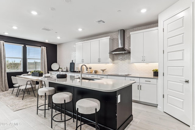 kitchen featuring sink, white cabinetry, black electric stovetop, wall chimney range hood, and a center island with sink