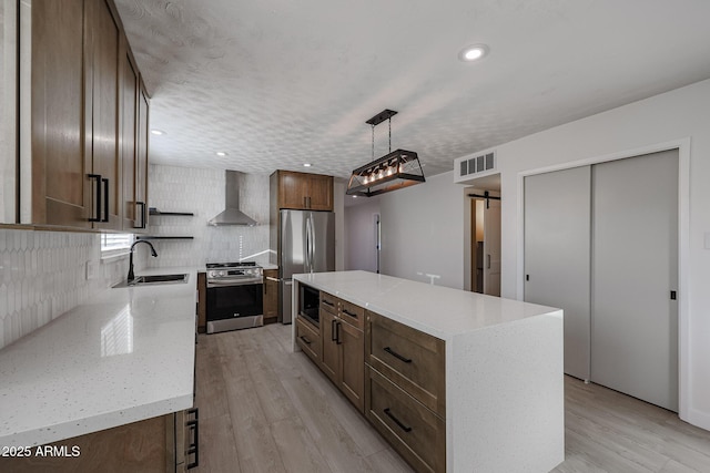 kitchen featuring wall chimney range hood, a barn door, appliances with stainless steel finishes, decorative light fixtures, and a kitchen island