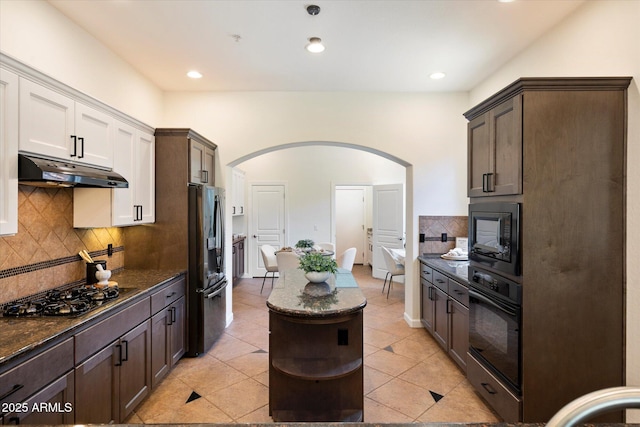 kitchen with decorative backsplash, arched walkways, black appliances, dark stone counters, and under cabinet range hood