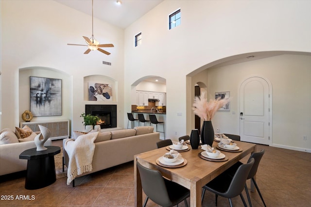 dining room with ceiling fan, arched walkways, high vaulted ceiling, a glass covered fireplace, and dark tile patterned floors