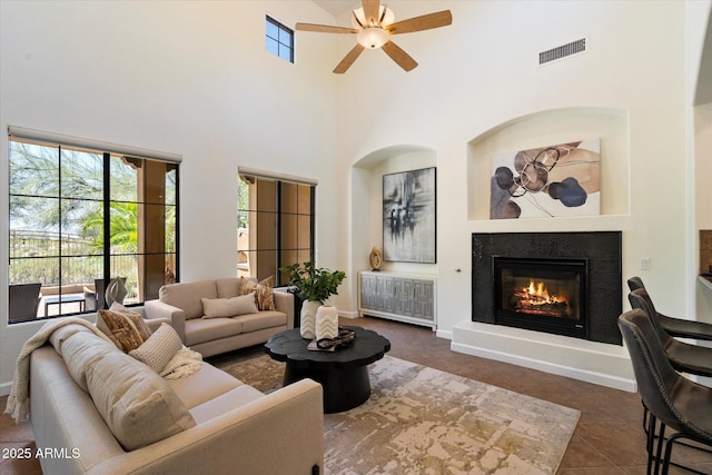 tiled living area with ceiling fan, visible vents, a towering ceiling, baseboards, and a glass covered fireplace