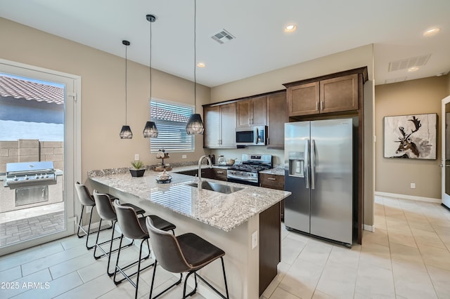 kitchen featuring stainless steel appliances, a sink, dark brown cabinets, light stone countertops, and a breakfast bar area