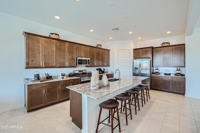 kitchen with appliances with stainless steel finishes, a sink, a kitchen bar, light stone counters, and a kitchen island with sink