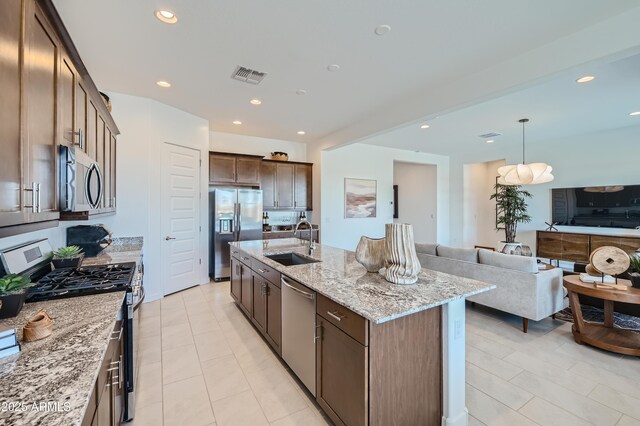 kitchen featuring appliances with stainless steel finishes, a sink, open floor plan, light stone counters, and an island with sink